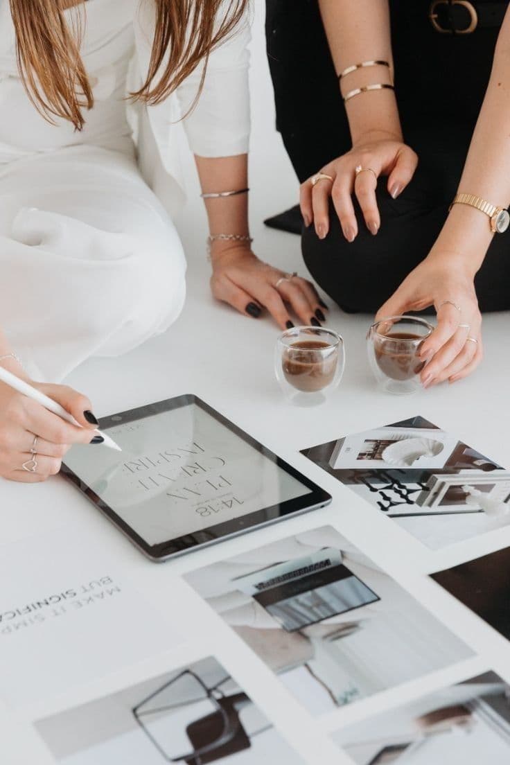 Two people kneeling at a white table, one drawing on a tablet among printed layouts