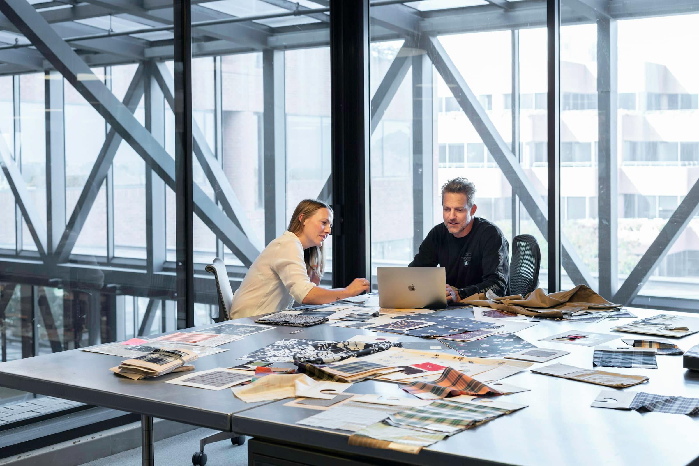 Two people at a long table spread with printed material, in a glass-walled room