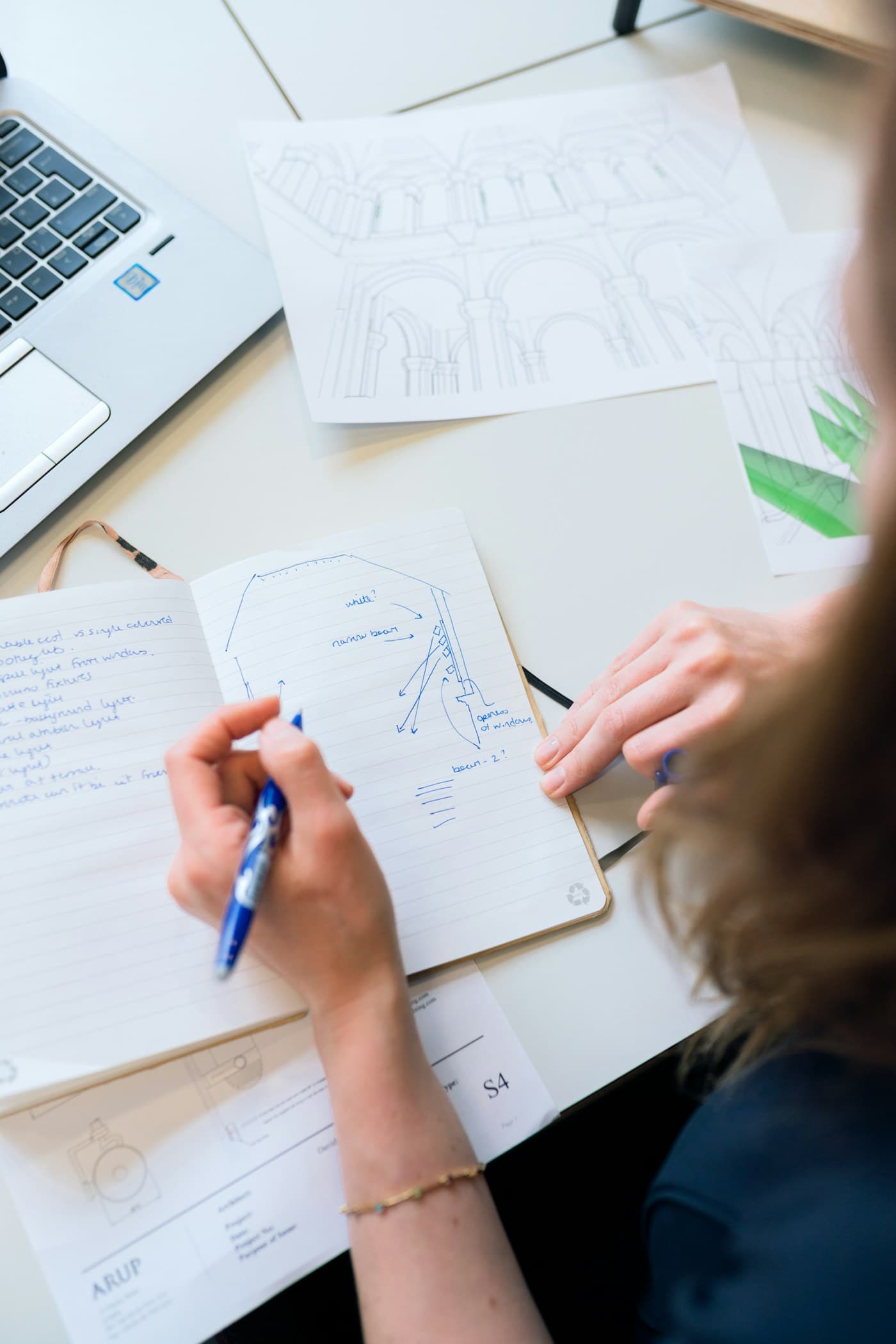 A hand writing notes in a notebook beside a sketched diagram and a laptop