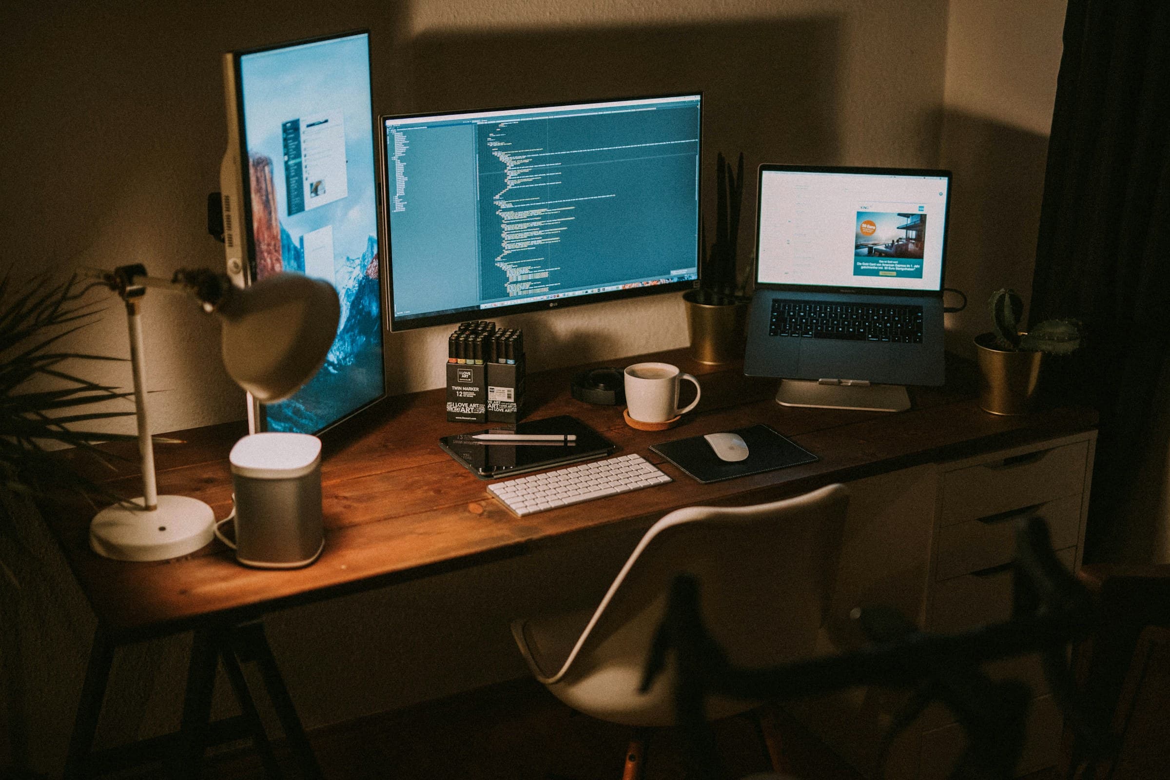 A lamp-lit desk with monitors showing code beside a laptop