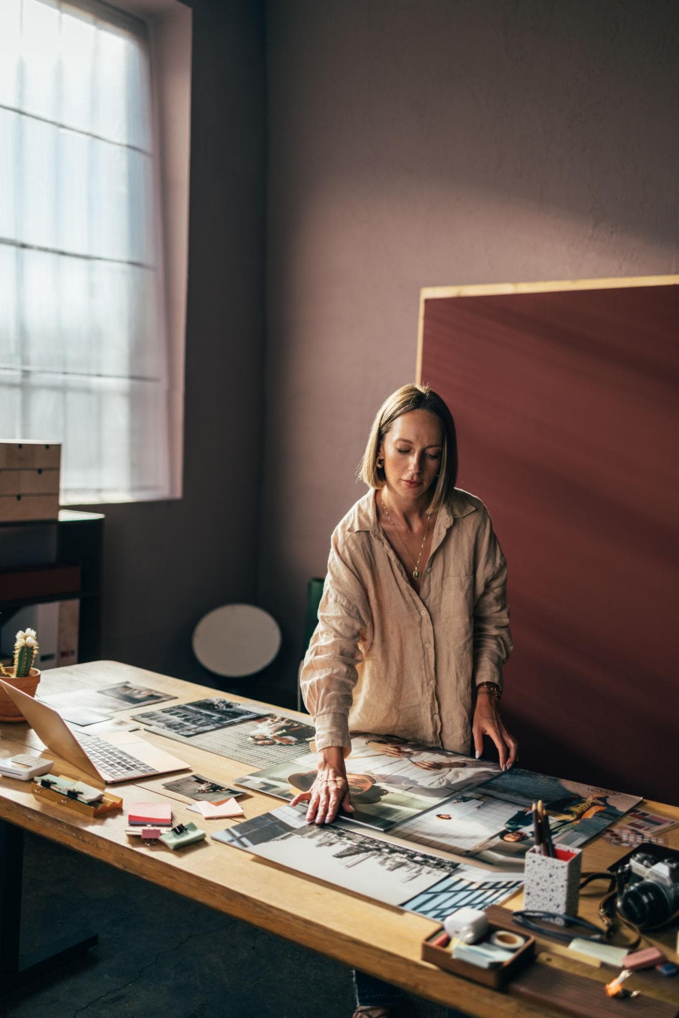 A woman standing at a long table covered in photographs and papers, in a room lit by a tall window