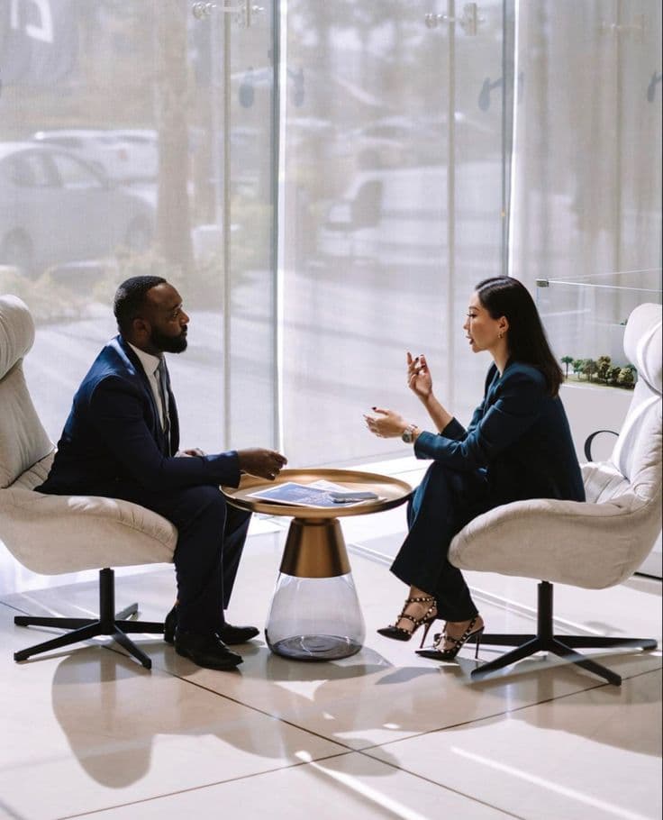 Two people in conversation in armchairs beside a tall window